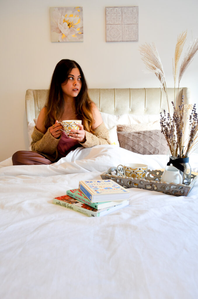 A photo of Lets Pour and ponder podcast host- Pooja. Pooja is sitting on a bed with a cup of coffee looking away from the camera