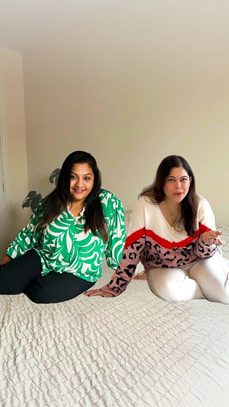 Nikita and Pooja podcast hosts of Lets Pour and Ponder, sit on a bed smiling at the camera.